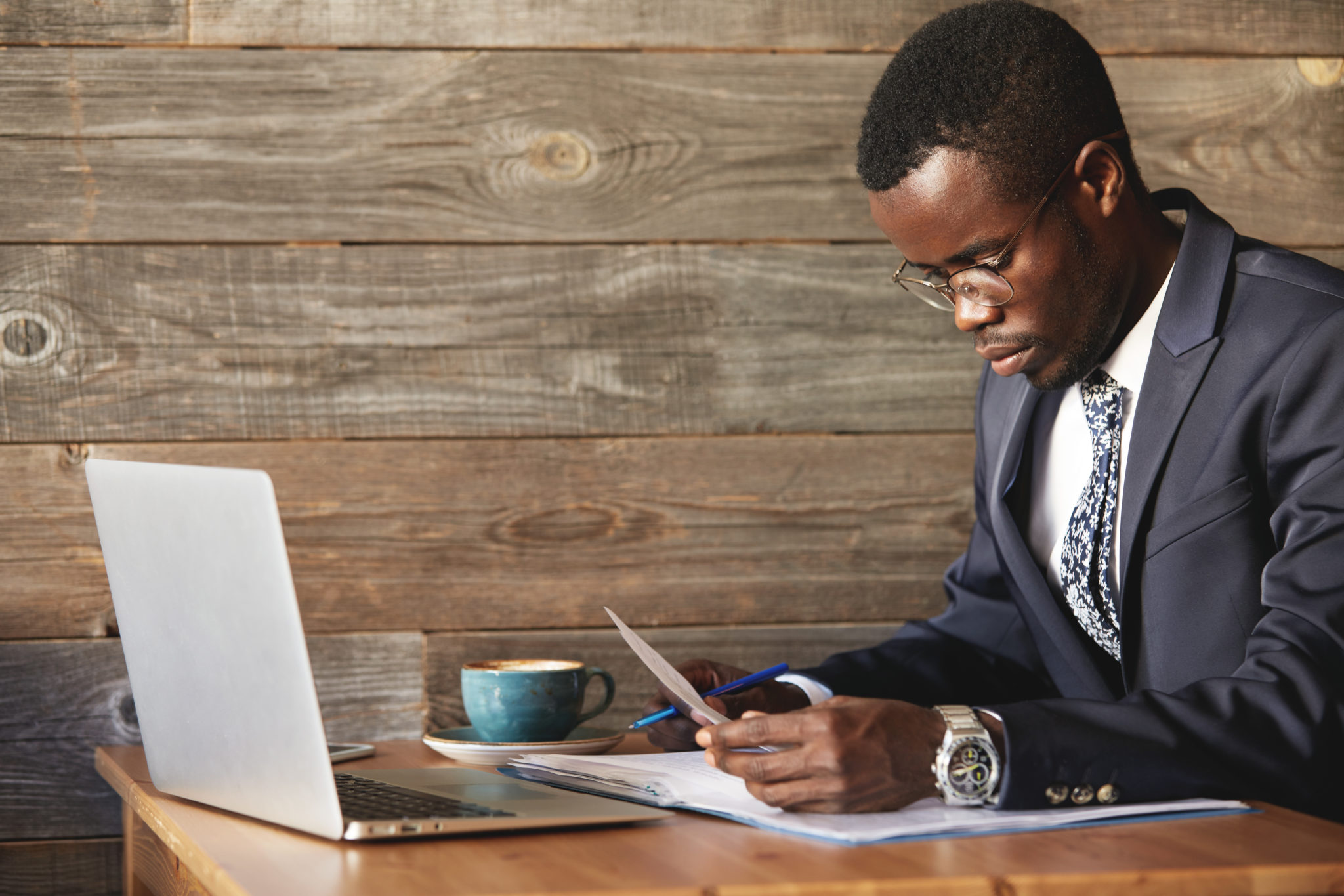 Young and successful businessman looking at contract and reading information in documents with blue pen in hand. Focused African man took laptop and digital devices with wi-fi for productive work. Image
