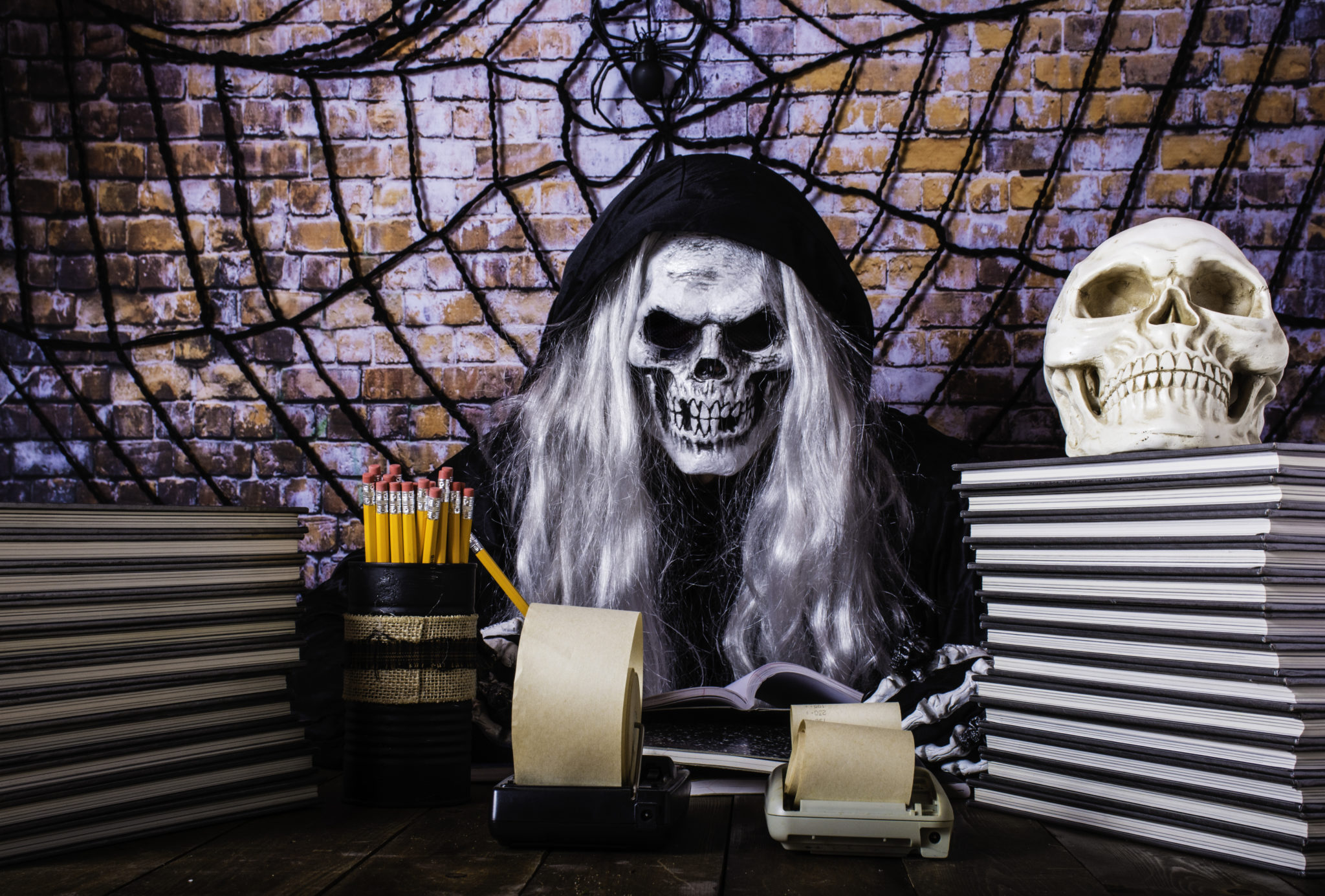 man in Grimm reaper robe scary skull mask long grey hair and skeleton hands counting lost souls with adding machine and pencils with brick wall background covered with large spider and web Image